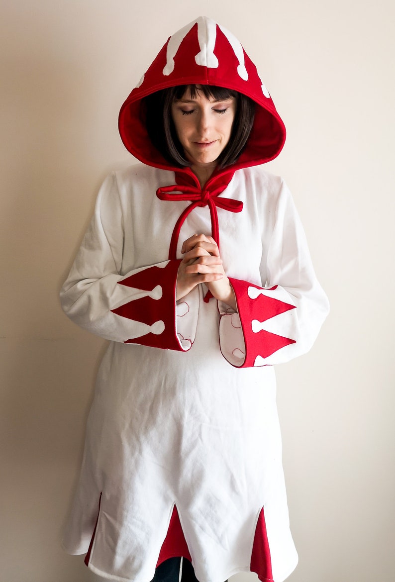 May include: A white hooded robe with red trim and white crown designs. The hood is tied with a red ribbon.