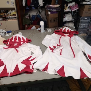 May include: Two white hooded sweatshirts with red accents. The sweatshirts have a pointed design on the bottom and sleeves. The hood has a red crown design.