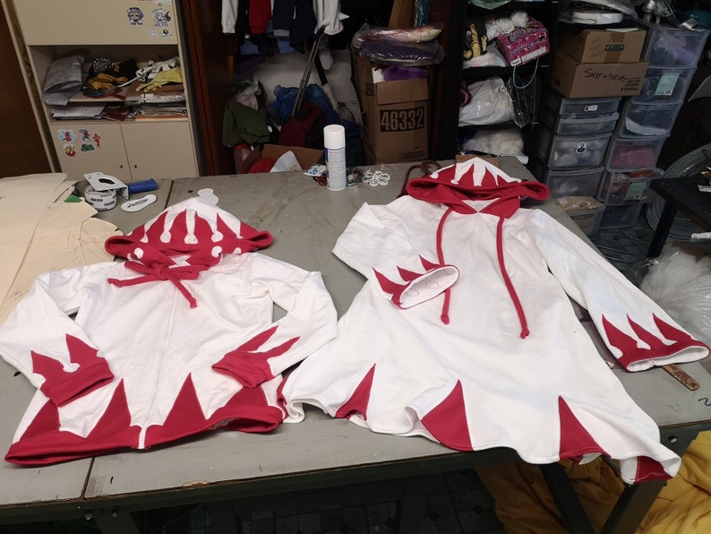 May include: Two white hooded sweatshirts with red accents. The sweatshirts have a pointed design on the bottom and sleeves. The hood has a red crown design.