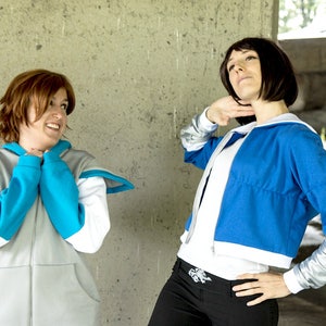May include: Two people wearing blue and white jackets with hoods. The person on the left is wearing a gray jacket with a blue hood and the person on the right is wearing a blue jacket with a white hood. They are standing in front of a concrete wall.