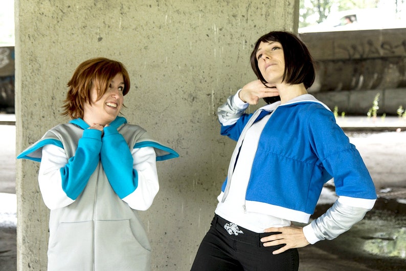 May include: Two people wearing blue and white jackets with hoods. The person on the left is wearing a gray jacket with a blue hood and the person on the right is wearing a blue jacket with a white hood. They are standing in front of a concrete wall.