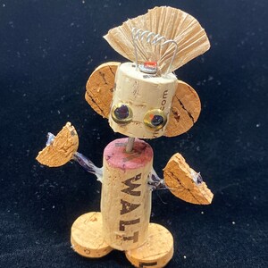 May include: A whimsical robot figurine made from wine corks, with a paper fan for a head, cork arms, and a cork body with the word "WALT" printed on it.