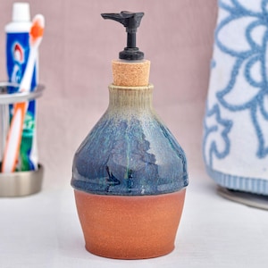 May include: A handmade ceramic soap dispenser with a dark blue and orange glaze. The dispenser has a black pump and a cork stopper. The background includes a toothbrush holder with toothbrushes and toothpaste, and a blue and white towel.