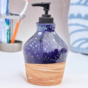 May include: A ceramic soap dispenser with a dark blue, speckled glaze on the upper portion and a light brown, wood-grain textured base. The dispenser has a black pump and is designed for bathroom use.