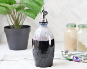 May include: A ceramic oil bottle with a stainless steel spout. The bottle is a dark blue at the base, fading to a lighter blue and white at the top. A small potted plant and jars are in the background.
