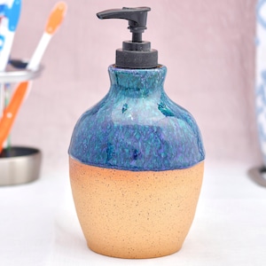 Puede incluir: Un dispensador de jabón de cerámica con una base beige moteada y una parte superior azul y verde brillante. El dispensador tiene una bomba negra, para uso en el baño.