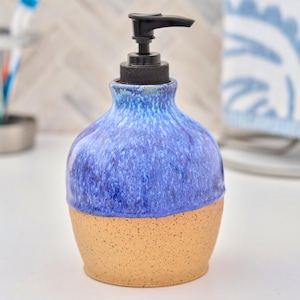 May include: A ceramic soap dispenser with a bulbous shape, featuring a glossy blue glaze on the upper portion and a speckled, unglazed tan base. The dispenser is topped with a functional black plastic pump mechanism for dispensing liquid soap.