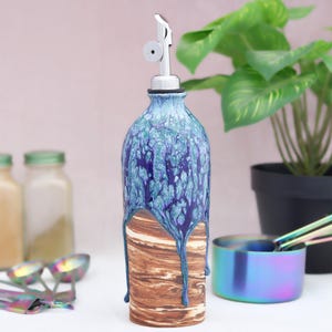 May include: A decorative ceramic oil bottle with a stainless steel spout. The bottle features a blue and purple glaze over a brown, wood-like base. Measuring spoons and a small bowl are in the foreground, with a potted plant in the background.