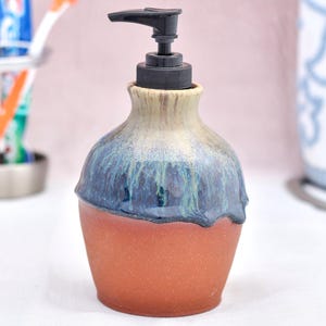 May include: A handmade ceramic soap dispenser with a black pump. The dispenser has a terracotta base, a blue and green glazed middle section, and a cream-coloured neck. The design is a unique addition to any bathroom.