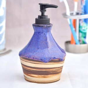 Puede incluir: Dispensador de jabón de cerámica hecho a mano con la parte superior esmaltada en azul y una base a rayas marrón y beige. El dispensador tiene una bomba negra y está diseñado para el baño.