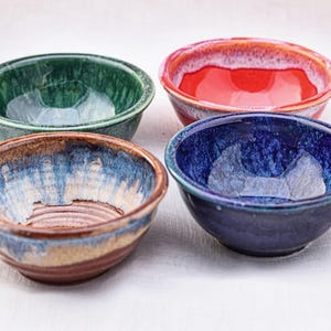 May include: Four small, handmade ceramic bowls in shades of green, red, blue, and brown. Each bowl has a unique glaze, creating a glossy, textured appearance. The bowls are arranged on a white surface, showcasing their vibrant colors and artisanal quality.
