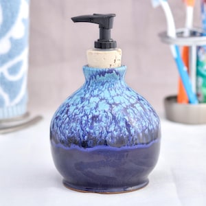Puede incluir: Un dispensador de jabón de cerámica con una base azul oscuro y una sección superior azul claro moteada. El dispensador tiene una bomba negra y un tapón de corcho. Una toalla azul y blanca y un porta cepillos de dientes están en el fondo.