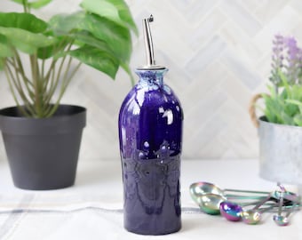 May include: A dark blue ceramic oil bottle with a stainless steel spout. The bottle has a glossy finish with a dripped glaze effect. The bottle is displayed with measuring spoons and potted plants.