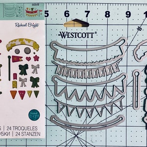 May include: A set of 24 metal dies for crafting, featuring a variety of banner and bow designs. The dies are arranged on a cutting mat with a grid pattern. The dies are silver in color and have a shiny finish. The packaging includes the text "THINLITS" and "Rachael Bright".