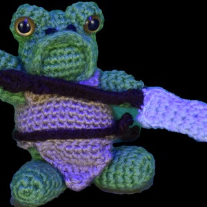 May include: A green crocheted frog wearing a purple and brown crocheted outfit and holding a light purple crocheted sword.