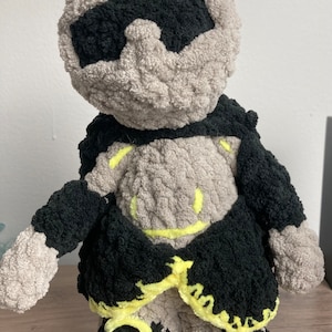 May include: A gray and black crocheted plush toy with a yellow mohawk and a black crocheted jacket. The toy has a yellow smiley face on its chest.