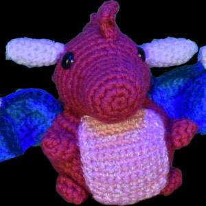 Puede incluir: Un pequeño juguete de dragón de crochet con cuerpo rosa y rojo, alas azules y detalles blancos.
