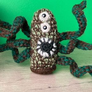 May include: A crocheted green and brown monster with multiple arms and large eyes. The monster has a white mouth with black teeth.