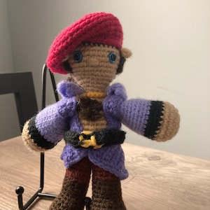 May include: Crocheted plush toy with a red beret, purple jacket, brown pants, and brown boots. The toy has blue eyes and a brown face.
