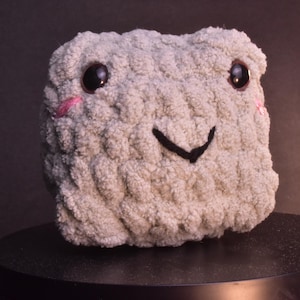 May include: A light gray, plush, square-shaped stuffed animal with black eyes, pink cheeks, and a black smile. The toy is made of a soft, textured material and sits on a black surface.