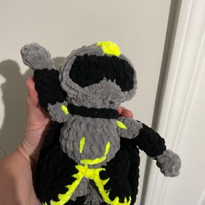 May include: A plush toy crafted from soft yarn, featuring a gray and black color scheme with vibrant neon yellow accents. The figure has a rounded head, a torso with neon yellow markings, and a black cape-like base with neon yellow trim.