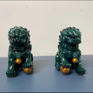 May include: Two jade green Foo Dog statues. Each statue is in a seated position, holding a gold ball under its paw. The statues have detailed manes and are adorned with gold bells around their necks. The background is a light gray.
