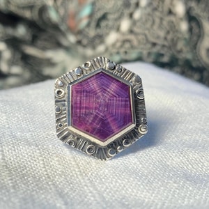May include: A silver ring featuring a hexagonal, purple gemstone. The gemstone has a patterned design and is set within a detailed, textured silver band. The ring's design includes small circular and linear embellishments around the gemstone.