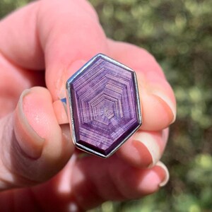 May include: A silver ring featuring a large, flat, hexagonal-shaped purple gemstone. The stone displays a distinct, concentric geometric pattern of nested hexagons in varying shades of deep violet and light purple, set within a smooth silver bezel.