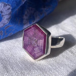 May include: A silver ring featuring a large, hexagonal, deep pink star sapphire gemstone. The stone displays a distinct six-rayed star pattern within its faceted surface, set in a polished silver band that encircles the finger.