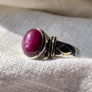 Op de afbeelding: Een ring met een grote, gladde, diepmagenta cabochon edelsteen gezet in een gepolijste zilveren band. De band bevat een goudkleurig accent bij de steen en een kleine, verzonken ruitvormige gouden inleg aan de zijkant van de metalen schacht.