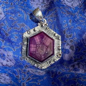 May include: A hexagonal pendant featuring a deep magenta star ruby gemstone set in a textured silver frame. The silver border displays a brushed finish with small raised spherical accents along the edges and a polished silver bail at the top.