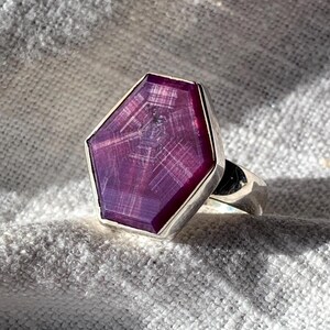 Puede incluir: Un anillo de plata con una gran gema púrpura facetada. La gema tiene una forma geométrica única, engastada en un bisel de plata. El anillo se muestra sobre un fondo de tela gris texturizada.