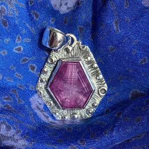 May include: A hexagonal pendant featuring a deep pink, faceted gemstone set in a textured silver frame with small spherical accents and a polished silver bail at the top, showcasing a distinct geometric design and metallic finish.