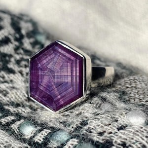 May include: A ring featuring a hexagonal, deep purple gemstone with a visible geometric, star-like pattern within the stone. The gemstone is set in a polished silver-colored metal band that extends from the back of the hexagonal setting.