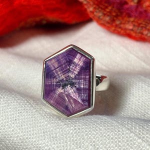 May include: A silver ring featuring a large, hexagonal-shaped purple gemstone with a distinct star-like pattern radiating from the center. The stone is set in a smooth, polished silver bezel that follows the six-sided geometry of the gem.