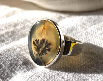 May include: A silver ring with an oval stone displaying a dendritic pattern, like a tree. The stone is a translucent yellowish-brown with dark brown and black inclusions. The ring has a polished silver band.