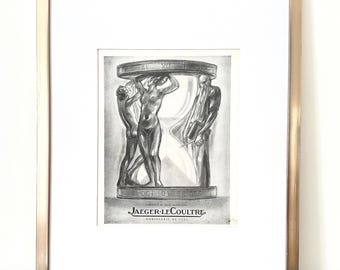 May include: Framed black and white print featuring an hourglass design with figures supporting the top. The print is in a gold-colored frame with a white mat. The text "JAEGER-LECOULTRE" is visible at the bottom. A pen rests below the frame.