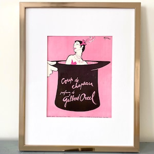 Puede incluir: Anuncio vintage enmarcado de perfume Gilbert Orcel. La obra de arte presenta a una mujer que emerge de un sombrero de copa, sobre un fondo rosa. El texto "Coup de chapeau parfums de Gilbert Orcel" se muestra en una elegante escritura.