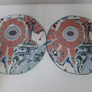 May include: Two white ceramic plates with a blue, green, red, and pink geometric design. The plates feature a central flower-like motif.