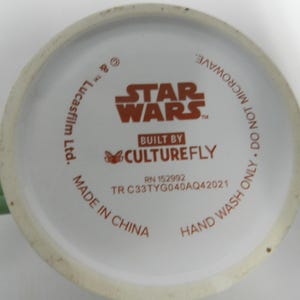 Op de afbeelding: Onderkant van een witte keramische mok met een groen handvat. De basis toont het "Star Wars"-logo in oranje, samen met tekst die aangeeft dat het is gebouwd door Culturefly en gemaakt in China. De tekst zegt ook "Alleen met de hand wassen. Niet in de magnetron."