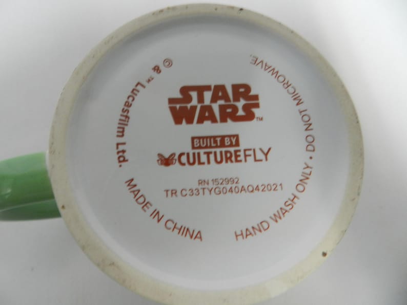Op de afbeelding: Onderkant van een witte keramische mok met een groen handvat. De basis toont het "Star Wars"-logo in oranje, samen met tekst die aangeeft dat het is gebouwd door Culturefly en gemaakt in China. De tekst zegt ook "Alleen met de hand wassen. Niet in de magnetron."