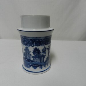 May include: A white and blue ceramic container with a cylindrical shape. The container features a blue and white Willow pattern design. The top portion is white, and the base has a slightly flared shape. The container is likely a decorative item or a small vase.