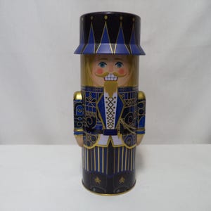 May include: A decorative tin nutcracker in a cylindrical shape. The nutcracker is painted with a detailed design featuring a blue and gold uniform, a blonde wig, and a smiling face. The hat is blue with gold trim.