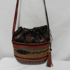 May include: A bucket-style shoulder bag featuring a dark, patterned fabric drawstring closure and a structured base with horizontal bands of red, brown, and black. The bag includes a long, thin shoulder strap and a decorative hanging tassel.