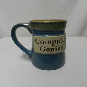 May include: A ceramic mug with a blue base and handle, a tan band with the words "Computer Genius", and a green rim. The mug is designed for hot beverages.