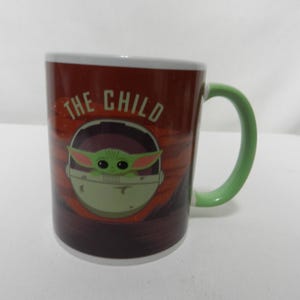 Op de afbeelding: Een keramische mok met een afbeelding van The Child, ook bekend als Baby Yoda, uit de Star Wars-serie. De mok heeft een rode achtergrond met de woorden "THE CHILD" erop gedrukt. Het handvat is groen en de binnenrand is wit.