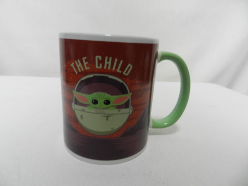 Op de afbeelding: Een keramische mok met een afbeelding van The Child, ook bekend als Baby Yoda, uit de Star Wars-serie. De mok heeft een rode achtergrond met de woorden "THE CHILD" erop gedrukt. Het handvat is groen en de binnenrand is wit.