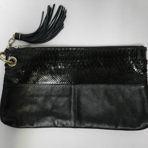 May include: Black clutch with a textured snakeskin-like upper section and a smooth leather lower section. A black tassel hangs from a gold-tone chain and ring. The bag is rectangular.