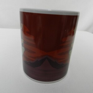 Op de afbeelding: Een keramische koffiemok met een donkerbruine achtergrond en een witte rand. De mok heeft een ontwerp met rode en witte accenten. De bodem van de mok heeft een reeks horizontale lijnen.