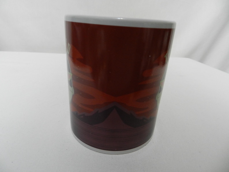 Op de afbeelding: Een keramische koffiemok met een donkerbruine achtergrond en een witte rand. De mok heeft een ontwerp met rode en witte accenten. De bodem van de mok heeft een reeks horizontale lijnen.